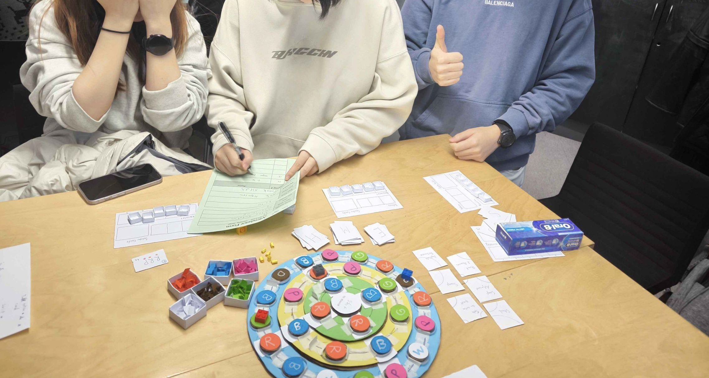 Two testers seated around the board mid-session, with Quest Cards and boxes of Colour Blocks on the table